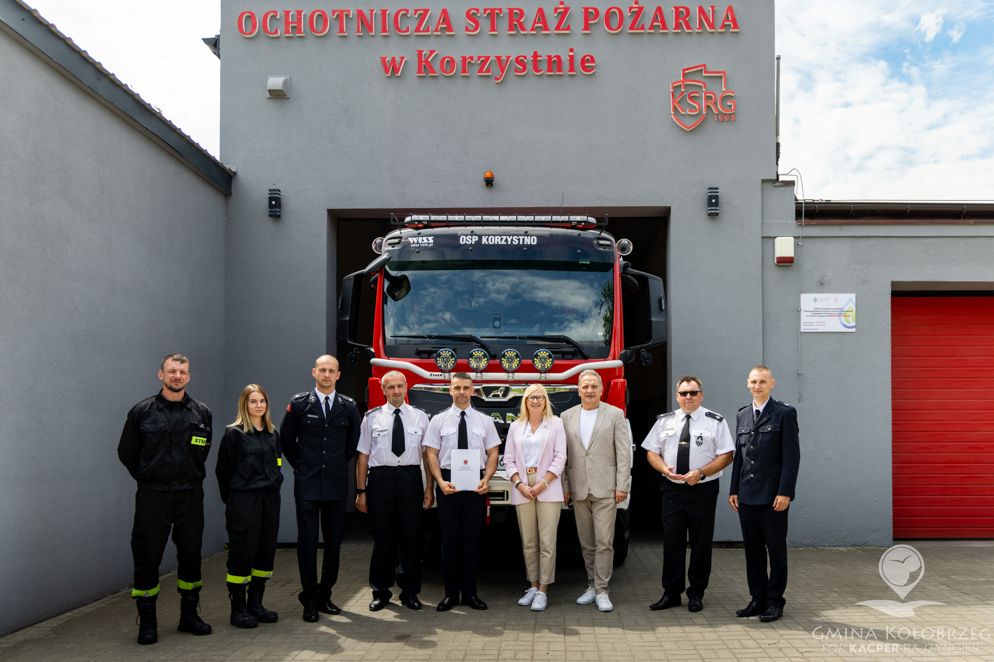 Zdjęcie przedstawiające grupę strażak&oacute;w oraz przedstawicieli samorządu pozujących przed remizą Ochotniczej Straży Pożarnej w Korzystnie. W tle widoczny jest w&oacute;z strażacki zaparkowany w garażu jednostki.