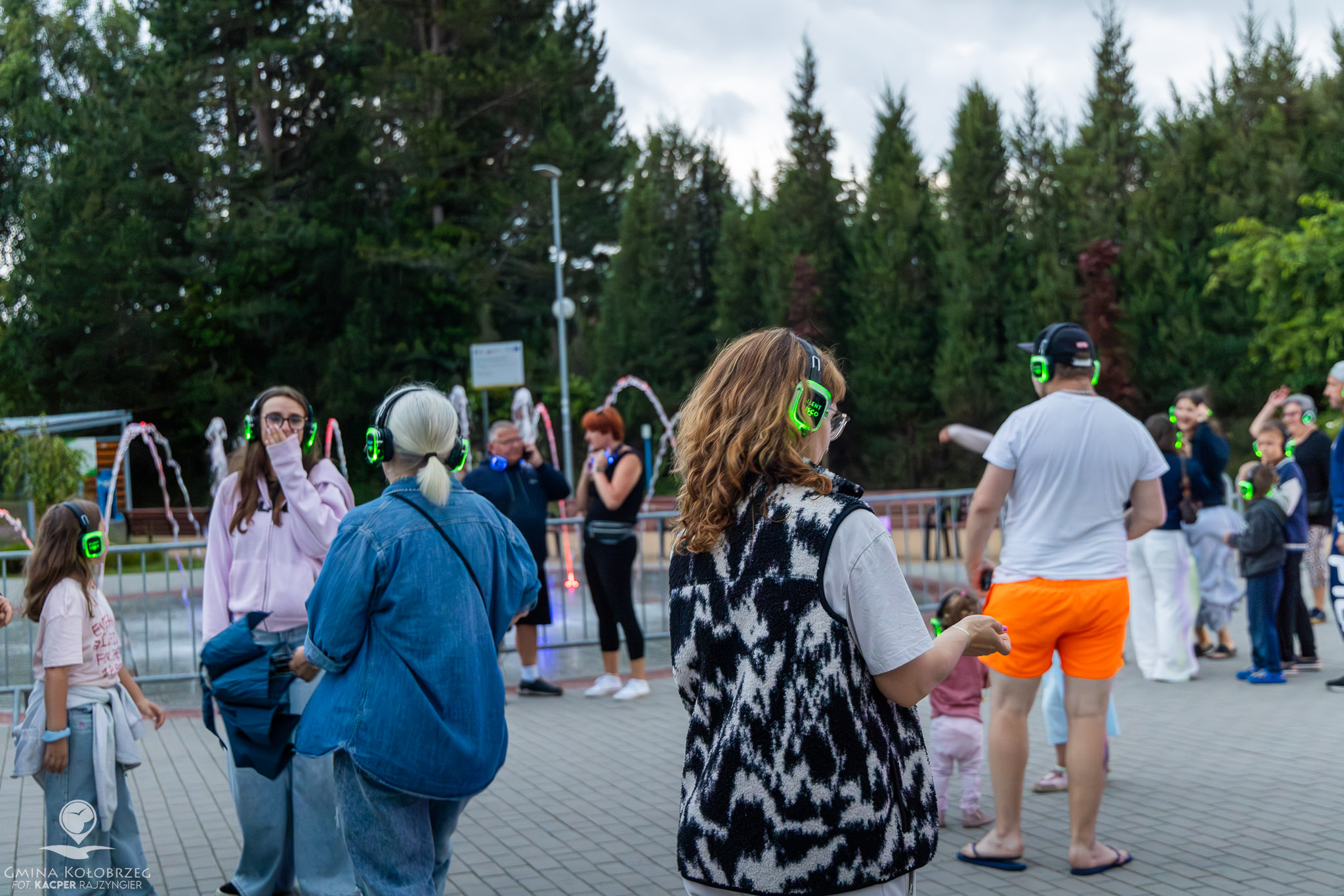 Ujęcie przedstawia uczestnik&oacute;w Silent Disco w słuchawkach, bawiących się na świeżym powietrzu przy podświetlonej fontannie. W tle widoczna jest zieleń oraz grupa os&oacute;b w r&oacute;żnym wieku korzystających z atrakcji.