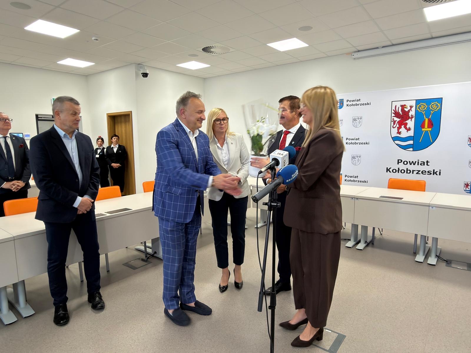 Zdjęcie przedstawia oficjalne spotkanie w sali Powiatu Kołobrzeskiego. Widać przekazanie kwiat&oacute;w przy mikrofonach, uczestnik&oacute;w wydarzenia oraz ściankę z herbem i napisem &bdquo;Powiat Kołobrzeski&rdquo;.